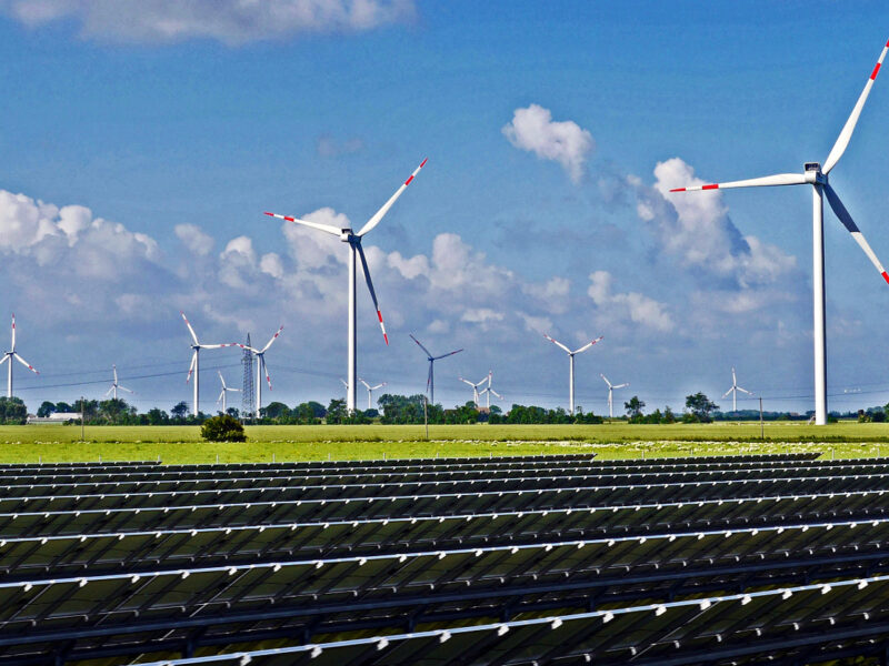 Solar panels, wind turbines, and an electricity pylon; unidentified location. Commons, by hpgruesen.